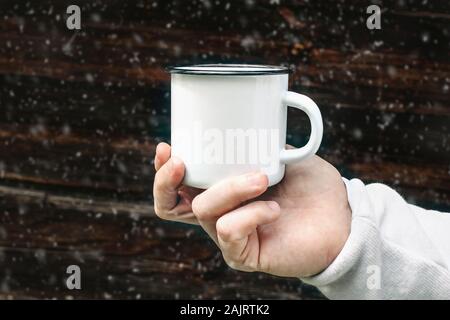 Close up of man voyageurs hand holding metal mug avec boisson chaude avec troubles de la neige. Thé, café en plein air vous détendre de temps. Maquette de la cuvette en émail blanc, Banque D'Images