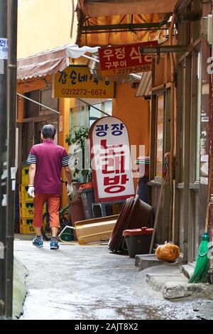 Un homme coréen d'âge moyen passe devant de petites boutiques le matin le long d'une ruelle latérale dans le marché de Gwangjang à Séoul, en Corée du Sud. Banque D'Images