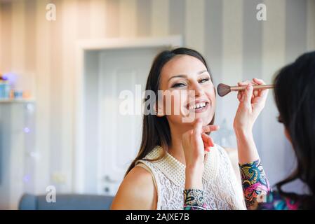 Jeune femme brune à la caméra en pendant la séance de maquillage où un artiste de maquillage professionnel effectue touche finale avec un pinceau doux Banque D'Images