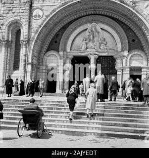 Années 1950, visiteurs à l'entrée de la Basilique Rosaire, Lourdes, France, une église catholique à la 'Sanctuaire de la Vierge De Lourdes' ou au domaine, une région de terre entourant le sanctuaire catholique (Grotto). Un lieu de pèlerinage pour beaucoup de la foi chrétienne, en particulier les malades et les infirmes, comme on peut le voir sur la photo par un homme dans un transport invalide de l'époque par les marches de l'église. Banque D'Images