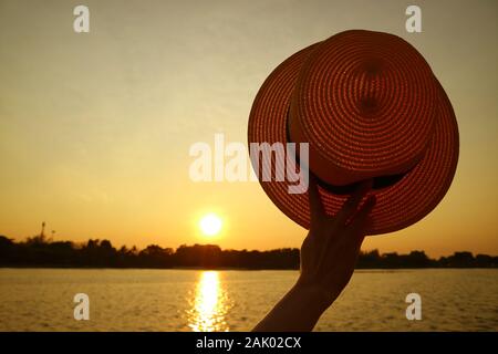 Woman's hand holding un chapeau de paille en montée contre sunset sky Banque D'Images