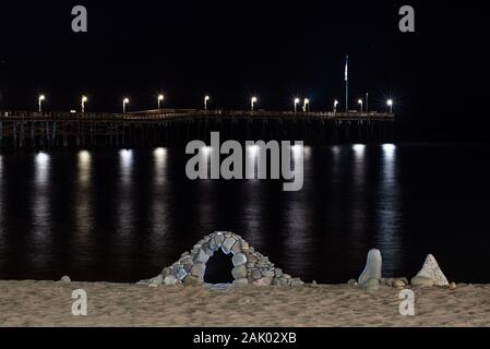 Ciel de nuit est sombre avant l'aube sur la plage de rochers empilés rétroéclairé délicatement dans un arc avec les lumières de jetée de Ventura brillants dans les lampes. Banque D'Images