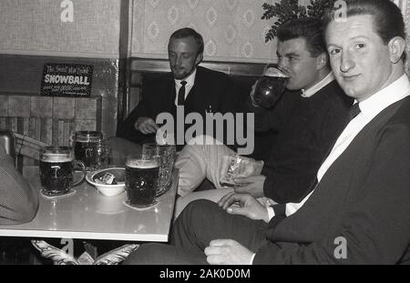 Années 1960, historiques, trois hommes appréciant la bière chopes de verre et chips de pommes de terre assis à une table dans un coin d'un bar ou pub, England, UK. Dans une publicité pour la nouvelle boisson "boule de neige" est sur le foyer de la cheminée. Banque D'Images