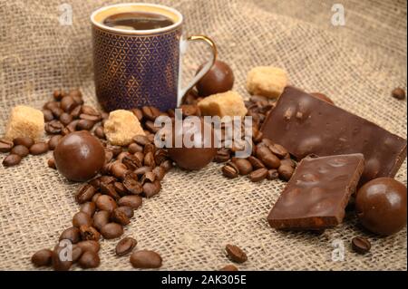 Une tasse de café, grains de café, des morceaux de sucre brun et de chocolat sur fond de tissu de fabrication locale. Close up Banque D'Images