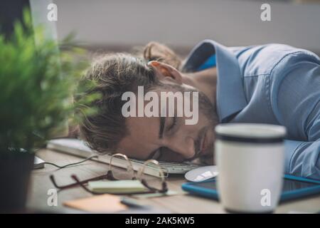S'est fatigué. Jeune homme barbu en chemise bleue ayant une sieste au travail Banque D'Images