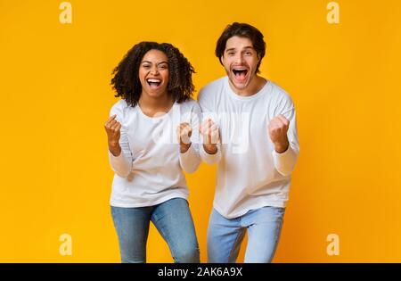 Grande Victoire. Cheeful race mixte couple celebrating success, élever les mains avec excitation sur fond jaune en studio, l'espace libre Banque D'Images