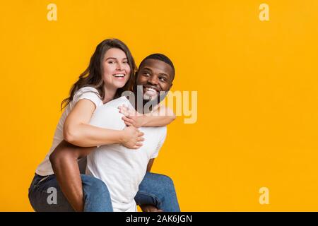 Heureux couple interracial. Cheerful black guy piggibacking sa copine joyeuse, de s'amuser ensemble sur fond jaune studio, l'espace vide Banque D'Images