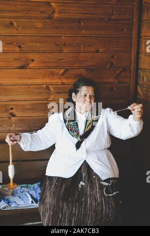 Santana, Madeira, Portugal - Sep 24, 2019 : Femme en costume traditionnel, assis sur un banc en bois et filage de la laine à la main. L'artisanat, l'artisanat ancien. Prises dans le parc à thème de Madère. Photo verticale. Banque D'Images