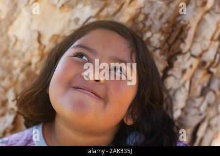 Une jeune fille aux joues potelées qui est plein de joie et de sourire tout en vous tenant à côté d'un arbre et profiter du soleil. Banque D'Images