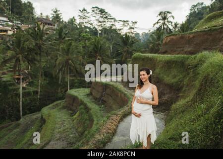 Jeune femme enceinte en robe blanche avec vue sur les rizières en terrasses de Bali dans la lumière du soleil du matin. Harmonie avec la nature. Concept de la grossesse. Banque D'Images