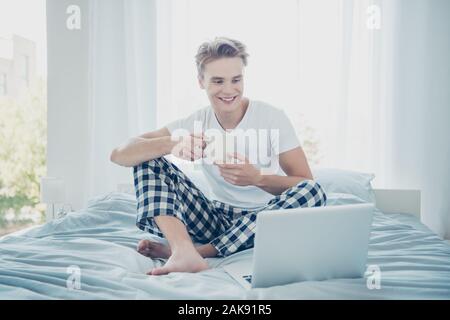 Photo pleine longueur de cheveux blonds gaie positive guy s'asseoir sur le lit tenir tasse avec boissons caféine utiliser son ordinateur portable pour regarder l'usure plaid à carreaux comédie Banque D'Images