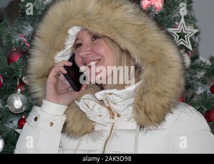 Young woman with fur hood using mobile phone in front of Christmas Tree Banque D'Images