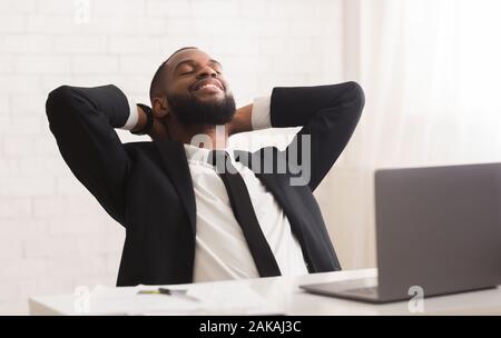 Happy afro businessman se reposant après une réunion fructueuse dans son bureau, se penchant en arrière avec les yeux fermés et le sourire sur son visage Banque D'Images