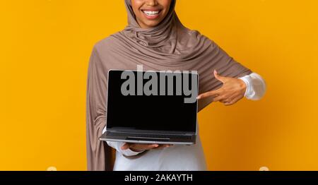 Ordinateur portable avec écran noir en blanc dans les mains de jeune fille musulmane afro méconnaissable en hijab, debout sur fond jaune, panorama avec de l'espace libre, récolte Banque D'Images