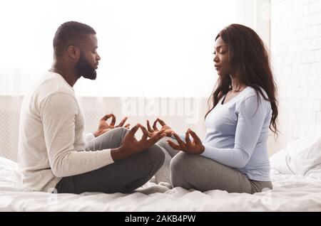 Le yoga prénatal. Femmes enceintes afro woman doing yoga avec son mari à la maison, assis sur le lit face à face avec les yeux fermé Banque D'Images