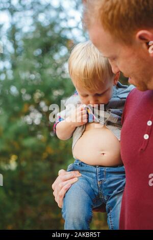 Un petit garçon dans les bras de papa soulève sa chemise pour vérifier son estomac Banque D'Images