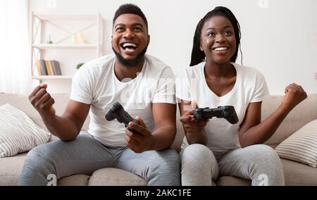 Passe-temps amusant pour les couples. Guy noir joyeux et célébrer la victoire en jeu vidéo, manettes, ce holding poings et riant, panorama Banque D'Images