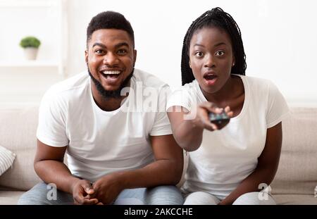 Programme tv choquant. Black couple surpris en regardant la télé avec bouche ouverte, passage d'une chaîne à l'appareil photo avec l'expression visage hébété Banque D'Images