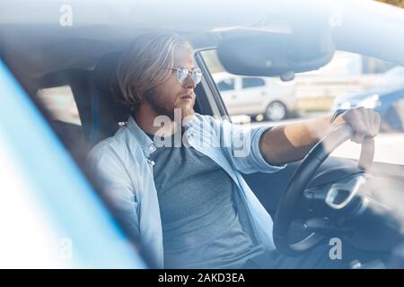 Le transport. Jeune homme barbu dans les verres voyageant en voiture électrique assis à l'intérieur du pare-brise vue à travers la conduite confiante Banque D'Images