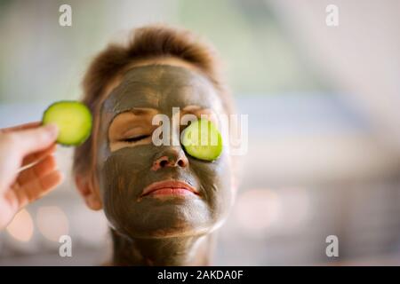 Mid-adult woman avec un masque de boue sur et tranches de concombre sur les yeux. Banque D'Images