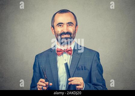 Coiffure barbu souriant portant veste élégante avec nœud Papillon rouge, qui posent avec ciseaux et peigne. Adultes élégant man studio portrait, isolée à gra Banque D'Images