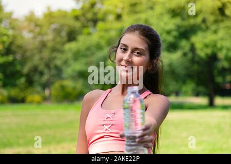 Souriante jeune adolescente dans le sport et l'eau potable à donner à un ami bouteille d'une bonne vie saine après l'exécution de l'exercice dans parc avec accueil chaleureux sunli Banque D'Images