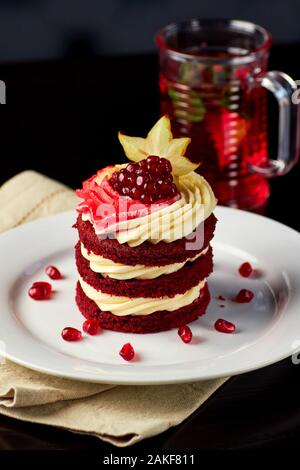 Délicieux gâteau de velours rouge décoré de crème fouettée, grenades et caramboles sur plaque blanche, servi avec un verre de thé sur la table en bois. Banque D'Images