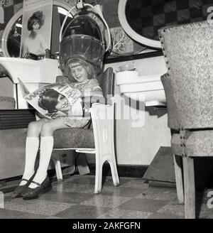 Années 1960, historiques, une jeune fille dans un salon de coiffure dames adultes, assis sur une chaise en lisant un magazine féminin, England, UK. Banque D'Images