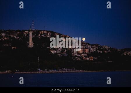 Trieste vue sur la ville de nuit avec la pleine lune s'élevant de la mer méditerranée. Célèbre tour lumineuse visible. Banque D'Images