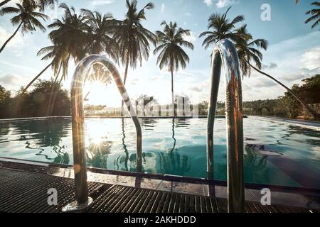 Détente en station touristique. Piscine au milieu des cocotiers contre lagoon au coucher du soleil. Banque D'Images