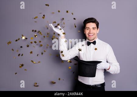 Portrait d'illusionniste positif tenue cylindre chapeau faire la mise au point épeler étoile dorée confetti voler chute dans l'air porter chemise blanc noeud noir isolé Banque D'Images