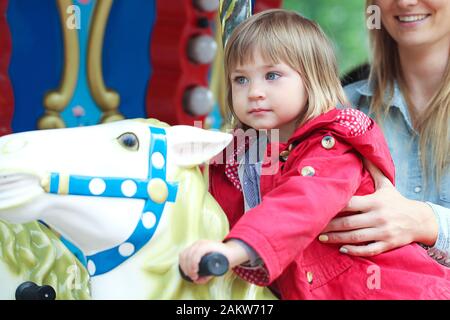 Cute little girl sitting on carousel horse. Sa mère debout à côté d'eux et maintenant Banque D'Images