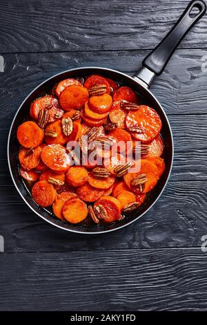 Pommes de terre douces confites avec sucre brun, sirop d'érable, jus d'orange et noix de pécan dans une poêle sur une table en bois noir, vue verticale de dessus, libre Banque D'Images