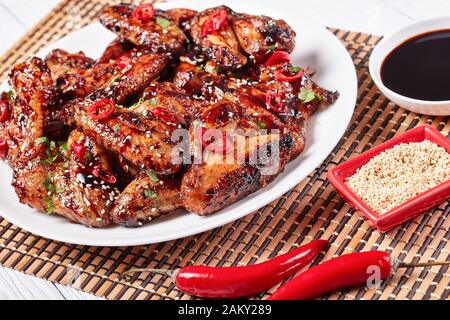 Gros plan d'ailes de poulet croustillantes de Teriyaki parsemées de graines de sésame, de morceaux de piment et de persil sur une plaque blanche sur un tapis de bambou, vue de l'ab Banque D'Images