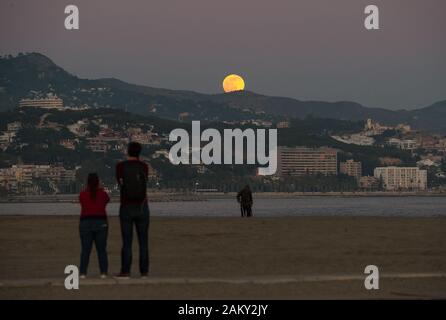 Les gens regardent la pleine lune (connu sous le nom de ce mois comme 'Wolf Moon') qu'il s'élève au-dessus de l'horizon à la plage pendant la première éclipse partielle de l'année. En cette année 2020 aura six éclipses, dont une éclipse solaire totale. Banque D'Images