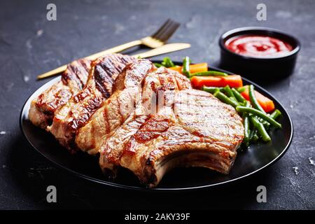 Gros plan de côtelettes de porc grillé servies avec une salade de haricots verts et de carottes sur une plaque noire sur une table en béton avec des couverts dorés Banque D'Images