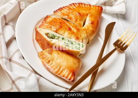 Calzone fraîchement cuit, pizza fermée avec garniture de épinards et de fromage servie sur une plaque blanche avec fourche dorée et couteau sur une table en bois, cuis italien Banque D'Images