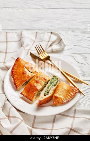 Calzone fraîchement cuit, pizza fermée avec garniture de épinards et de fromage servie sur une plaque blanche avec fourche dorée et couteau sur une table en bois, cuis italien Banque D'Images