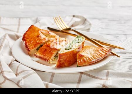 Calzone fraîchement cuit, pizza fermée avec garniture de épinards et de fromage servie sur une plaque blanche avec fourche dorée et couteau sur une table en bois, cuis italien Banque D'Images