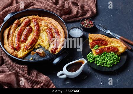 Gros plan d'une partie de Toad dans le trou, Saucisse Toad, saucisses dans le Yorkshire pudding sur une plaque noire servie avec des petits pois et de la sauce à l'oignon, Banque D'Images