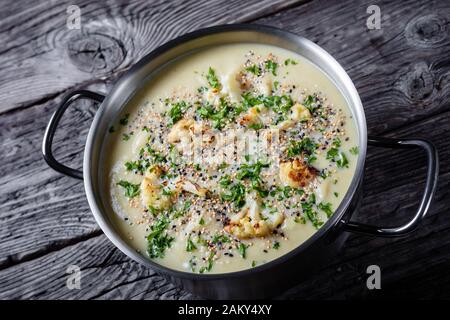 Gros plan de la soupe de chou-fleur à l'ail rôti crémeux avec du fromage asiago et une garniture de quinoa et de graines de chia croustillante dans une casserole sur un woo rustique Banque D'Images