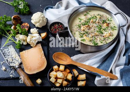 Gros plan de la soupe de Chou-Fleur à l'ail rôtie d'Asiago dans un marmite avec ingrédients et croûtons sur une table en béton, vue horizontale d'en haut Banque D'Images