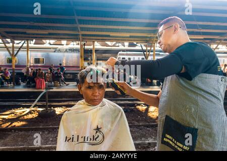 Bangkok/Thaïlande-décembre 2019: un barbier de rue donnant la coupe libre à son client masculin sur une plate-forme de train à la gare de Hua Lamphong. Banque D'Images