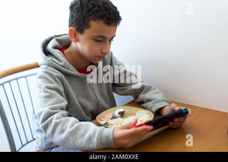 garçon mange mal distrait par le jeu. Enfants mangeant du riz et surfer sur Internet ou jouer à des jeux vidéo sur la console Banque D'Images