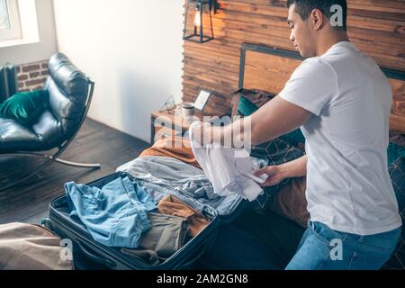 Jeune homme aux cheveux noirs en blanc tshirt étant occupé avec des vêtements d'emballage Banque D'Images