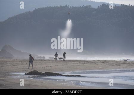 Les pompiers hélicoptère transportant un seau d'eau au-dessus d'une plage où peu de gens à pied avec les chiens Banque D'Images
