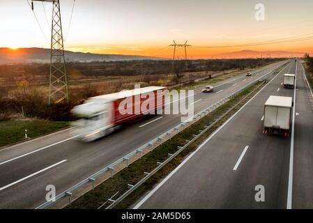 Camions et voitures de livraison en conduite à grande vitesse sur une route à travers le paysage rural. Mouvement flou rapide sur l'autoroute. Scène de fret sur le mois Banque D'Images