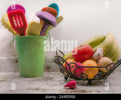 Comptoir de cuisine: Un seau de fantaisie contenant des outils de cuisine colorés et un bol de fruits en métal contenant des fruits et des légumes. Banque D'Images