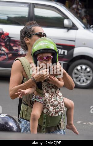 Jeune mère touristique plaçant un casque de scooter vert citron sur son verre de soleil rose portant une jolie petite fille avant de monter sur le scooter. Banque D'Images