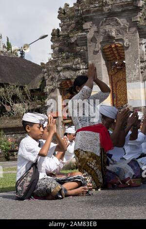 Jeune garçon balinais, hindou vêtu de sarong coloré, chemise blanche, bandeau blanc assis sur le sol du temple avec des mains claquées dans la prière au-dessus de sa tête Banque D'Images